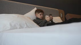 Το αγόρι βρίσκεται στο κρεβάτι με ένα smartphone στα χέρια του Ανακοίνωση και παιχνίδια σχετικά με το smartphone απόθεμα βίντεο Το αγόρι βρίσκεται στο κρεβάτι με ένα smartphone στα χέρια του Ανακοίνωση και παιχνίδια σχετικά με το smartphone απόθεμα βίντεο