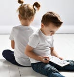 Το αγόρι και το κορίτσι δύο παιδιών στις άσπρα μπλούζες και το τζιν παντελόνι κάθονται το ένα κοντά στο άλλο και παίζουν το θωρηκ στοκ εικόνες Το αγόρι και το κορίτσι δύο παιδιών στις άσπρα μπλούζες και το τζιν παντελόνι κάθονται το ένα κοντά στο άλλο και παίζουν το θωρηκ στοκ εικόνες