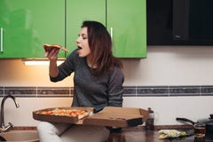 Голодная девушка сидя на кухне и есть вкусную пиццу стоковые изображения rf Голодная девушка сидя на кухне и есть вкусную пиццу стоковые изображения rf