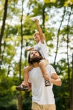 Идти в мальчика леса белокурый одел в белой футболке сидя на плечах его бородатого отца и держит стоковое изображение Идти в мальчика леса белокурый одел в белой футболке сидя на плечах его бородатого отца и держит стоковое изображение