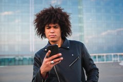 Красивый молодой человек с афро волосами используя умный телефон и выглядеть серьезный, предпосылка города стоковое изображение rf Красивый молодой человек с афро волосами используя умный телефон и выглядеть серьезный, предпосылка города стоковое изображение rf