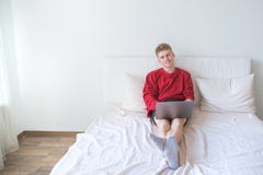 Молодой человек в красной фуфайке сидит на кровати в светлой комнате и работах на ноутбуке и взглядах на камере стоковое изображение Молодой человек в красной фуфайке сидит на кровати в светлой комнате и работах на ноутбуке и взглядах на камере стоковое изображение