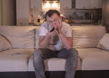 Молодой человек с пробуренным взглядом дома в вечере сидит на софе с дистанционным управлением и смотрит ТВ стоковые фотографии rf Молодой человек с пробуренным взглядом дома в вечере сидит на софе с дистанционным управлением и смотрит ТВ стоковые фотографии rf