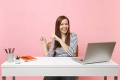 Молодая смеясь женщина в случайных одеждах указывает указательный палец в сторону сидит, работа на белом столе с современным ноут стоковая фотография Молодая смеясь женщина в случайных одеждах указывает указательный палец в сторону сидит, работа на белом столе с современным ноут стоковая фотография