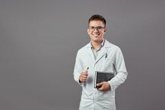 Умный усмехаясь парень в стеклах одетых в белом пальто держит тетрадь и лоток на серой предпосылке стоковые фотографии rf Умный усмехаясь парень в стеклах одетых в белом пальто держит тетрадь и лоток на серой предпосылке стоковые фотографии rf