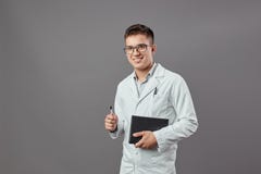 Умный усмехаясь парень в стеклах одетых в белом пальто держит тетрадь и лоток на серой предпосылке стоковое фото Умный усмехаясь парень в стеклах одетых в белом пальто держит тетрадь и лоток на серой предпосылке стоковое фото