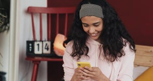 Ładna młoda kobieta ono uśmiecha się podczas gdy gawędzący na żółtym smartphone obsiadaniu na łóżku w domu Dziewczyna wysyła wiad zbiory Ładna młoda kobieta ono uśmiecha się podczas gdy gawędzący na żółtym smartphone obsiadaniu na łóżku w domu Dziewczyna wysyła wiad zbiory