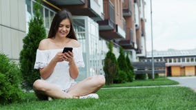 Śliczna dziewczyna używa telefon komórkowego podczas gdy siedzący na trawie blisko hotelu, gawędzi z przyjaciółmi zbiory wideo Śliczna dziewczyna używa telefon komórkowego podczas gdy siedzący na trawie blisko hotelu, gawędzi z przyjaciółmi zbiory wideo