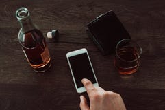 Ένα άτομο πιέζει ένα δάχτυλο σε ένα κινητό τηλέφωνο Έπειτα στον πίνακα είναι ένα ποτήρι του ουίσκυ, ένα μπουκάλι του ουίσκυ και έ στοκ εικόνες Ένα άτομο πιέζει ένα δάχτυλο σε ένα κινητό τηλέφωνο Έπειτα στον πίνακα είναι ένα ποτήρι του ουίσκυ, ένα μπουκάλι του ουίσκυ και έ στοκ εικόνες
