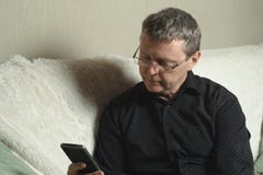 Ένα κουρασμένο άτομο σε ένα μαύρο πουκάμισο εξετάζει την οθόνη ενός smartphone καθμένος σε έναν καναπέ στοκ φωτογραφίες με δικαίωμα ελεύθερης χρήσης Ένα κουρασμένο άτομο σε ένα μαύρο πουκάμισο εξετάζει την οθόνη ενός smartphone καθμένος σε έναν καναπέ στοκ φωτογραφίες με δικαίωμα ελεύθερης χρήσης