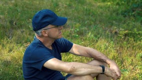Ένα λεπτό, σοβαρό γκρίζος-μαλλιαρό άτομο σε μια μπλε μπλούζα, η ΚΑΠ και τα γυαλιά κάθονται στην πράσινη χλόη στο δάσος σε ένα καλ απόθεμα βίντεο Ένα λεπτό, σοβαρό γκρίζος-μαλλιαρό άτομο σε μια μπλε μπλούζα, η ΚΑΠ και τα γυαλιά κάθονται στην πράσινη χλόη στο δάσος σε ένα καλ απόθεμα βίντεο