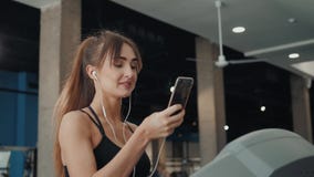 Αρκετά νέος αθλητής γυναικών με το smartphone που τρέχει treadmill στη γυμναστική, μουσική ακούσματος κοριτσιών γυναικών στο κινη απόθεμα βίντεο Αρκετά νέος αθλητής γυναικών με το smartphone που τρέχει treadmill στη γυμναστική, μουσική ακούσματος κοριτσιών γυναικών στο κινη απόθεμα βίντεο