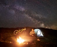 Γυναίκα που στηρίζεται τη νύχτα να στρατοπεδεύσει κοντά στην πυρά προσκόπων, σκηνή τουριστών, ποδήλατο κάτω από το σύνολο ουρανού στοκ εικόνες με δικαίωμα ελεύθερης χρήσης Γυναίκα που στηρίζεται τη νύχτα να στρατοπεδεύσει κοντά στην πυρά προσκόπων, σκηνή τουριστών, ποδήλατο κάτω από το σύνολο ουρανού στοκ εικόνες με δικαίωμα ελεύθερης χρήσης