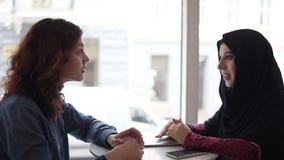 Δύο διεθνείς θηλυκοί φίλοι που μιλούν στον καφέ: η νέα μουσουλμανική γυναίκα στο μαύρο hijab μιλά στο θηλυκό της καυκάσιο φιλμ μικρού μήκους Δύο διεθνείς θηλυκοί φίλοι που μιλούν στον καφέ: η νέα μουσουλμανική γυναίκα στο μαύρο hijab μιλά στο θηλυκό της καυκάσιο φιλμ μικρού μήκους