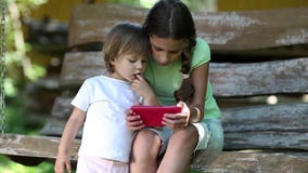 Δύο συμπαθητικά κορίτσια με το κόκκινο smartphone κάθονται στον πάγκο ταλάντευσης στον κήπο φιλμ μικρού μήκους Δύο συμπαθητικά κορίτσια με το κόκκινο smartphone κάθονται στον πάγκο ταλάντευσης στον κήπο φιλμ μικρού μήκους