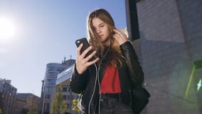 Ελκυστική νέα γυναίκα με μακρυμάλλη στα ακουστικά που χρησιμοποιούν το smartphone περπατώντας στο αστικό υπόβαθρο οδών απόθεμα βίντεο Ελκυστική νέα γυναίκα με μακρυμάλλη στα ακουστικά που χρησιμοποιούν το smartphone περπατώντας στο αστικό υπόβαθρο οδών απόθεμα βίντεο