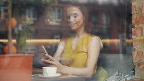 Ελκυστική νέα κυρία που χρησιμοποιεί το smartphone στη χαλάρωση καφέδων με τη συσκευή και τον καφέ απόθεμα βίντεο Ελκυστική νέα κυρία που χρησιμοποιεί το smartphone στη χαλάρωση καφέδων με τη συσκευή και τον καφέ απόθεμα βίντεο
