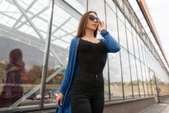 Η αρκετά προκλητική νέα γυναίκα hipster στη μοντέρνη μαύρη κορυφή σε ένα πλεκτό ακρωτήριο στα καθιερώνοντα τη μόδα τζιν στα σκοτε στοκ εικόνα Η αρκετά προκλητική νέα γυναίκα hipster στη μοντέρνη μαύρη κορυφή σε ένα πλεκτό ακρωτήριο στα καθιερώνοντα τη μόδα τζιν στα σκοτε στοκ εικόνα