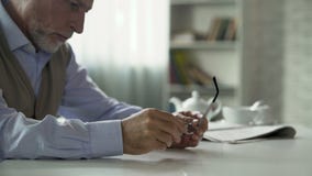 Η ηλικιωμένη αρσενική συνεδρίαση στον πίνακα και το τρίψιμο των χεριών, που βγάζουν τα γυαλιά, ανησύχησαν φιλμ μικρού μήκους Η ηλικιωμένη αρσενική συνεδρίαση στον πίνακα και το τρίψιμο των χεριών, που βγάζουν τα γυαλιά, ανησύχησαν φιλμ μικρού μήκους