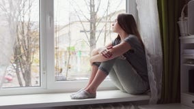 Η λυπημένη γυναίκα κάθεται στο windowsill, φαίνεται έξω το παράθυρο και χαμηλώνει το κεφάλι στα γόνατα φιλμ μικρού μήκους Η λυπημένη γυναίκα κάθεται στο windowsill, φαίνεται έξω το παράθυρο και χαμηλώνει το κεφάλι στα γόνατα φιλμ μικρού μήκους