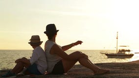 Η μητέρα και ο γιος κάθονται στην παραλία στο ηλιοβασίλεμα προσέχοντας ένα σκάφος περνούν απόθεμα βίντεο Η μητέρα και ο γιος κάθονται στην παραλία στο ηλιοβασίλεμα προσέχοντας ένα σκάφος περνούν απόθεμα βίντεο