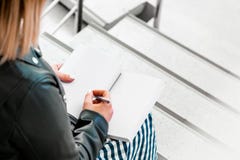 Η νέα γυναίκα κάνει τις σημειώσεις στο ιώδες σημειωματάριο στη βιβλιοθήκη και τη συνεδρίαση στα βήματα σκαλών στοκ φωτογραφίες Η νέα γυναίκα κάνει τις σημειώσεις στο ιώδες σημειωματάριο στη βιβλιοθήκη και τη συνεδρίαση στα βήματα σκαλών στοκ φωτογραφίες
