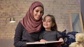 Η νέα γυναίκα στο hijab που χαμογελά και κάθεται στον καναπέ με την κόρη της και την διδάσκει πώς να διαβάσει, να κρατήσει, ευτυχ φιλμ μικρού μήκους Η νέα γυναίκα στο hijab που χαμογελά και κάθεται στον καναπέ με την κόρη της και την διδάσκει πώς να διαβάσει, να κρατήσει, ευτυχ φιλμ μικρού μήκους