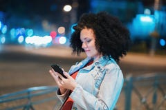 Η νέα όμορφη γυναίκα με την πολύ σγουρή τρίχα afro που χρησιμοποιεί το κινητό τηλέφωνο φώτισε τη νύχτα την οδό Ασυνήθιστο καθιερώ στοκ φωτογραφίες με δικαίωμα ελεύθερης χρήσης Η νέα όμορφη γυναίκα με την πολύ σγουρή τρίχα afro που χρησιμοποιεί το κινητό τηλέφωνο φώτισε τη νύχτα την οδό Ασυνήθιστο καθιερώ στοκ φωτογραφίες με δικαίωμα ελεύθερης χρήσης