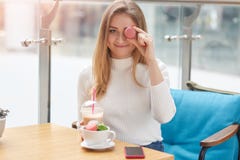 Η νέα όμορφη κυρία στο άσπρο πουκάμισο κάθεται στον πίνακα στον μπλε καναπέ και καλύπτει το μάτι της με ρόδινο macaroon χαρωπά εξ στοκ φωτογραφία με δικαίωμα ελεύθερης χρήσης Η νέα όμορφη κυρία στο άσπρο πουκάμισο κάθεται στον πίνακα στον μπλε καναπέ και καλύπτει το μάτι της με ρόδινο macaroon χαρωπά εξ στοκ φωτογραφία με δικαίωμα ελεύθερης χρήσης
