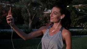 Η όμορφη γυναίκα με τα ακουστικά χτυπά στο τηλέφωνο μπροστά από ένα πάρκο απόθεμα βίντεο Η όμορφη γυναίκα με τα ακουστικά χτυπά στο τηλέφωνο μπροστά από ένα πάρκο απόθεμα βίντεο