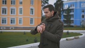 Κλείστε επάνω του νέου γενειοφόρου ατόμου στο καφετί παλτό που περπατά στην οδό κοντά στα κτήρια και που ελέγχει το ρολόι του Το απόθεμα βίντεο Κλείστε επάνω του νέου γενειοφόρου ατόμου στο καφετί παλτό που περπατά στην οδό κοντά στα κτήρια και που ελέγχει το ρολόι του Το απόθεμα βίντεο