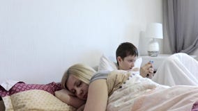 Κουρασμένη μητέρα γυναικών που ξυπνούν μετά από τον ύπνο και τη συνεδρίαση ήλιών της στο κρεβάτι και παίζοντας κινητά παιχνίδια σ απόθεμα βίντεο Κουρασμένη μητέρα γυναικών που ξυπνούν μετά από τον ύπνο και τη συνεδρίαση ήλιών της στο κρεβάτι και παίζοντας κινητά παιχνίδια σ απόθεμα βίντεο