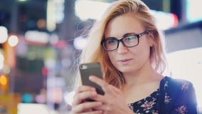 Μια νέα γυναίκα στα γυαλιά χρησιμοποιεί ένα smartphone σε μια πόλη νύχτας Στα πλαίσια των μουτζουρωμένων φω'των της νύχτας στοκ εικόνα Μια νέα γυναίκα στα γυαλιά χρησιμοποιεί ένα smartphone σε μια πόλη νύχτας Στα πλαίσια των μουτζουρωμένων φω'των της νύχτας στοκ εικόνα