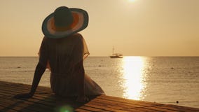 Μια νέα γυναίκα συναντά την ανατολή στην αποβάθρα Κάθεται και εξετάζει τον ήλιο και το σκάφος στη θάλασσα Όνειρα και ειδύλλιο στοκ εικόνα Μια νέα γυναίκα συναντά την ανατολή στην αποβάθρα Κάθεται και εξετάζει τον ήλιο και το σκάφος στη θάλασσα Όνειρα και ειδύλλιο στοκ εικόνα