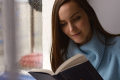 Νέα γυναίκα brunette στο μπλε μάλλινο πουλόβερ, που διαβάζει ένα ενδιαφέρον βιβλίο στο windowsill στο δωμάτιο στη βροχή, νεφελώδη Στοκ εικόνες με δικαίωμα ελεύθερης χρήσης Νέα γυναίκα brunette στο μπλε μάλλινο πουλόβερ, που διαβάζει ένα ενδιαφέρον βιβλίο στο windowsill στο δωμάτιο στη βροχή, νεφελώδη Στοκ εικόνες με δικαίωμα ελεύθερης χρήσης