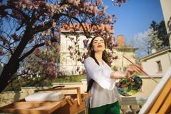 Νέα εκμετάλλευση καλλιτεχνών γυναικών brunette στα χέρια μια βούρτσα και μια παλέτα Κοντά σε την το δέντρο magnolia και ο διάφορο στοκ εικόνα Νέα εκμετάλλευση καλλιτεχνών γυναικών brunette στα χέρια μια βούρτσα και μια παλέτα Κοντά σε την το δέντρο magnolia και ο διάφορο στοκ εικόνα