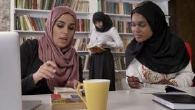 Νέες μουσουλμανικές γυναίκες στο hijab που εξηγεί κάτι στις μαύρες γυναίκες στο hijab, που μελετά στη βιβλιοθήκη και που προετοιμ απόθεμα βίντεο Νέες μουσουλμανικές γυναίκες στο hijab που εξηγεί κάτι στις μαύρες γυναίκες στο hijab, που μελετά στη βιβλιοθήκη και που προετοιμ απόθεμα βίντεο