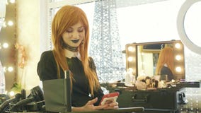 Νέο όμορφο κορίτσι με αποκριές makeup που χρησιμοποιούν το έξυπνο τηλέφωνο στο σαλόνι ομορφιάς απόθεμα βίντεο Νέο όμορφο κορίτσι με αποκριές makeup που χρησιμοποιούν το έξυπνο τηλέφωνο στο σαλόνι ομορφιάς απόθεμα βίντεο