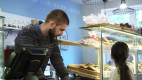 Νέος αρσενικός αρτοποιός που εργάζεται στο κατάστημά του που βοηθά το μικρό κορίτσι που επιλέγει τα επιδόρπια απόθεμα βίντεο Νέος αρσενικός αρτοποιός που εργάζεται στο κατάστημά του που βοηθά το μικρό κορίτσι που επιλέγει τα επιδόρπια απόθεμα βίντεο