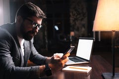 Νέος γενειοφόρος επιχειρηματίας που χρησιμοποιεί το τηλέφωνο καθμένος από τον ξύλινο πίνακα στο σύγχρονο γραφείο τη νύχτα Άνθρωπο Στοκ εικόνα με δικαίωμα ελεύθερης χρήσης Νέος γενειοφόρος επιχειρηματίας που χρησιμοποιεί το τηλέφωνο καθμένος από τον ξύλινο πίνακα στο σύγχρονο γραφείο τη νύχτα Άνθρωπο Στοκ εικόνα με δικαίωμα ελεύθερης χρήσης