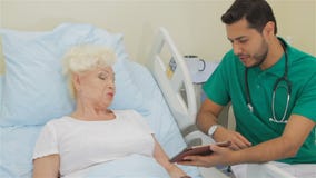 Ο γιατρός παρουσιάζει κάτι στην ταμπλέτα του στο θηλυκό ασθενή απόθεμα βίντεο Ο γιατρός παρουσιάζει κάτι στην ταμπλέτα του στο θηλυκό ασθενή απόθεμα βίντεο