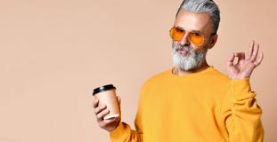 Ο μοντέρνος πρεσβύτερος ατόμων που εξετάζει τη κάμερα, που έχει την ΚΑΠ με τον καφέ στα όπλα και παρουσιάζει ένα σημάδι εντάξει μ στοκ εικόνα με δικαίωμα ελεύθερης χρήσης Ο μοντέρνος πρεσβύτερος ατόμων που εξετάζει τη κάμερα, που έχει την ΚΑΠ με τον καφέ στα όπλα και παρουσιάζει ένα σημάδι εντάξει μ στοκ εικόνα με δικαίωμα ελεύθερης χρήσης