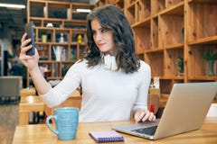 Ο νέος όμορφος σπουδαστής κοριτσιών blogger με τη μακριά σγουρή τρίχα σε ένα άσπρο πουλόβερ και με τα ακουστικά στο λαιμό κάνει έ στοκ εικόνα με δικαίωμα ελεύθερης χρήσης Ο νέος όμορφος σπουδαστής κοριτσιών blogger με τη μακριά σγουρή τρίχα σε ένα άσπρο πουλόβερ και με τα ακουστικά στο λαιμό κάνει έ στοκ εικόνα με δικαίωμα ελεύθερης χρήσης