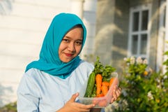 Πορτρέτο της ελκυστικής νέας γυναίκας hijab που κρατά ένα σύνολο κάδων των λαχανικών μπροστά από το σπίτι της στοκ φωτογραφία με δικαίωμα ελεύθερης χρήσης Πορτρέτο της ελκυστικής νέας γυναίκας hijab που κρατά ένα σύνολο κάδων των λαχανικών μπροστά από το σπίτι της στοκ φωτογραφία με δικαίωμα ελεύθερης χρήσης