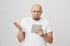 Πορτρέτο της ταραγμένης και ανίδεης φαλακρής γενειοφόρου αρσενικής πρότυπης ταμπλέτας και εκμετάλλευσης με ένα άλλο χέρι κοιτάζον στοκ εικόνες Πορτρέτο της ταραγμένης και ανίδεης φαλακρής γενειοφόρου αρσενικής πρότυπης ταμπλέτας και εκμετάλλευσης με ένα άλλο χέρι κοιτάζον στοκ εικόνες