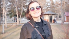 Πορτρέτο της χαμογελώντας καυκάσιας νέας γυναίκας στα γυαλιά ηλίου σε ένα πάρκο πόλεων o φιλμ μικρού μήκους Πορτρέτο της χαμογελώντας καυκάσιας νέας γυναίκας στα γυαλιά ηλίου σε ένα πάρκο πόλεων o φιλμ μικρού μήκους