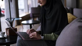 Συγκεντρωμένη νέα μουσουλμανική γυναίκα που εργάζεται στο σύγχρονο lap-top στον καφέ Ελκυστική γυναίκα στο hijab που κρατά το lap φιλμ μικρού μήκους Συγκεντρωμένη νέα μουσουλμανική γυναίκα που εργάζεται στο σύγχρονο lap-top στον καφέ Ελκυστική γυναίκα στο hijab που κρατά το lap φιλμ μικρού μήκους