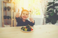 Το ευτυχές κορίτσι παιδιών τρώει τα λαχανικά και παρουσίαση αντίχειρων στοκ εικόνα Το ευτυχές κορίτσι παιδιών τρώει τα λαχανικά και παρουσίαση αντίχειρων στοκ εικόνα