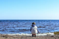 Το μικρό κορίτσι σε μια εγκαταλειμμένη παραλία κάθεται και συλλέγει τις πέτρες και τα κοχύλια ενάντια στο μπλε ουρανό και τα όμορ στοκ εικόνα Το μικρό κορίτσι σε μια εγκαταλειμμένη παραλία κάθεται και συλλέγει τις πέτρες και τα κοχύλια ενάντια στο μπλε ουρανό και τα όμορ στοκ εικόνα