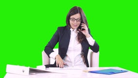 Το νέο brunette σε ένα πράσινο υπόβαθρο κάθεται πίσω από ένα γραφείο στην αρχή και που μιλά στο τηλέφωνο Στον πίνακα είναι έγγραφ φιλμ μικρού μήκους Το νέο brunette σε ένα πράσινο υπόβαθρο κάθεται πίσω από ένα γραφείο στην αρχή και που μιλά στο τηλέφωνο Στον πίνακα είναι έγγραφ φιλμ μικρού μήκους